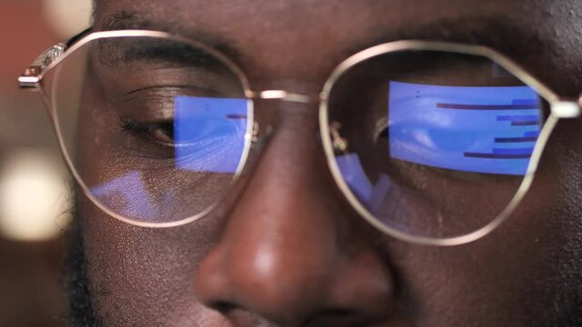 Close-up of graphs, charts and diagrams reflected in glasses of focused african american businessman analyzing data on computer screen. Dark-skinned man in eyeglasses watching presentation on laptop