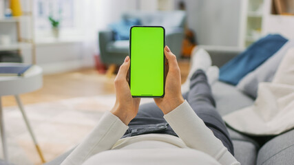 Young Woman at Home Uses Green Mock-up Screen Smartphone. She's Sitting On a Couch in His Cozy Living Room. Point of View Shot.