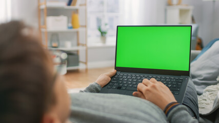 Man at Home Laying on a Couch Using with Green Mock-up Screen Laptop Computer. Guy Using Laptop Device, Browsing Internet, Watching Content, Videos.