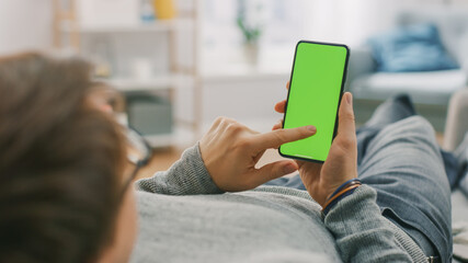 Man at Home Lying on a Couch using Smartphone with Green Mock-up Screen, Doing Swiping, Scrolling...