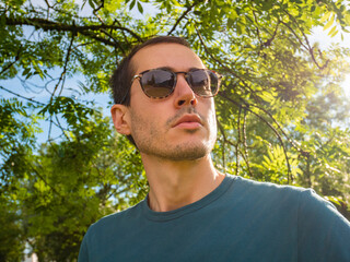 Portrait of a young man wears sunglasses. Green trees of a forest on the background. Sun rays beat on the glasses, polarized lens concept.