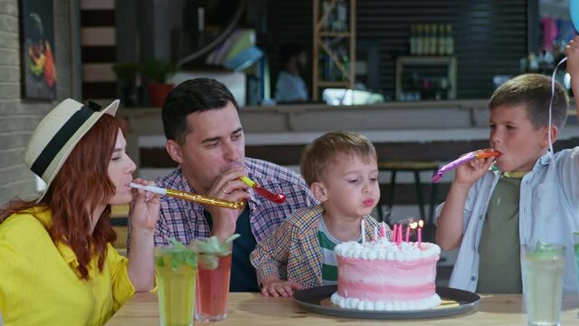 happy party, little cute male child with his loving parents and brother with balloons and pipes celebrates his birthday blows out candles on delicious cake and bites festive dessert