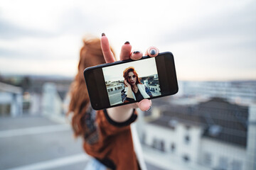 Young beautiful woman holding smart phone in outstretched hand. Phone displaying her portrait. Red haired female model.