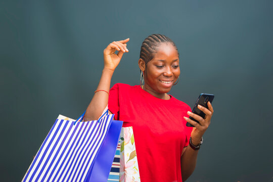 a young beautiful african lady wearing red cloth isolated over grey background feeling excited about what she saw on her cellphone with holding a shopping bag