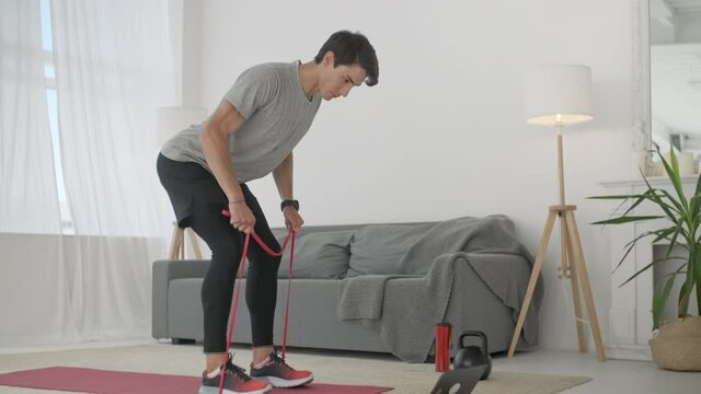 Handsome Young Man in a Gray T-Shirt and Black Shorts Does Sports at Home Online. Active fitness male showing exercise for back and arms muscle using rubber band
