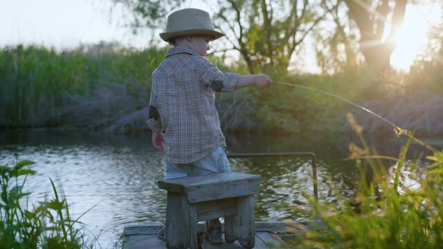 child in nature, cute beautiful baby in hat and stick in hands has fun on pier, playing in pond among trees against sunset at weekend into countryside