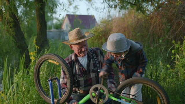 lifestyle, happy elderly man and charming ginny grandson in hats paddle spend time spinning wheels and pedals of bicycle outdoors during family outdoor recreation amon trees and grass