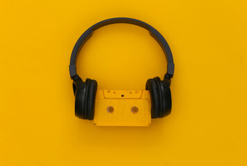 Black stereo headphones with a retro audio cassette on a yellow background. Top view