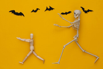 Halloween scary concept. Wooden puppet runs away from the skeleton on yellow background with flying...