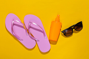 Beach vacation, travel concept. Sunblock bottle and sunglasses, flip flops on yellow bright sunny background. Top view. Flat lay