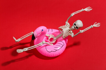 Toy skeleton with inflatable flamingo on red bright background. Halloween theme. Beach vacation concept. Summer rest. © splitov27