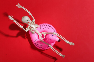 Toy skeleton with inflatable flamingo on red bright background. Halloween theme. Beach vacation concept. Summer rest. © splitov27