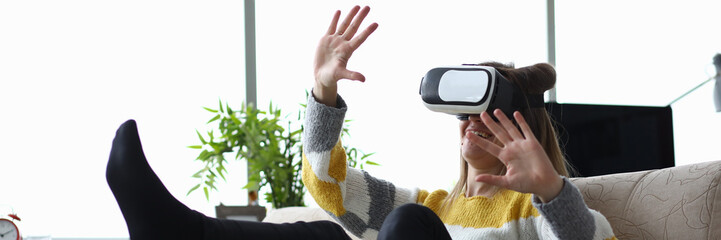 Close-up of young woman touch something using modern virtual reality glasses. Pretty female sitting on sofa in funny pose. Vr experience and entertainment concept