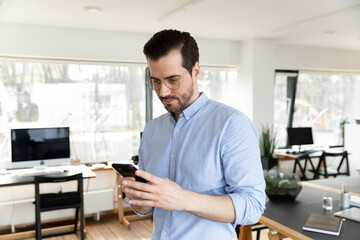 Young Caucasian male employee in glasses look on cellphone screen text or message in office, concentrated businessman use modern smartphone gadget, browse wireless internet at workplace