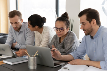 Serious diverse young businesspeople sit at desk in office cooperate brainstorm using laptops, concentrated multiethnic colleagues employees work in groups on computer at meeting, teamwork concept