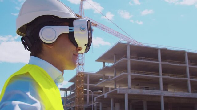Professional builder in VR helmet standing in front of construction site and using virtual and augmented reality technologies. Office building and crane background. Real estate and investment.