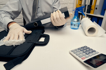 concept of cleaning or disinfecting the office desk - a businessman cleans the workplace, computer keyboard, document folders, uses a spray gun sanitizer, gloves and paper napkins.