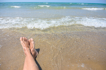 Feet of a man on a sandy beach, in the background a sea surf and horizon