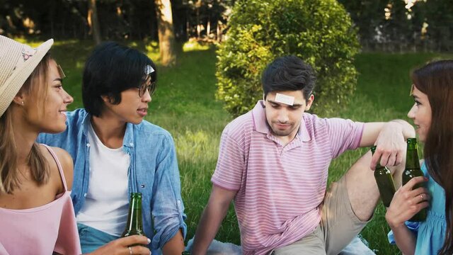 Friends playing guessing game who am I with stickers on forehead. They talking, laughing, drinking beer, sitting on green grass in park. Slow motion