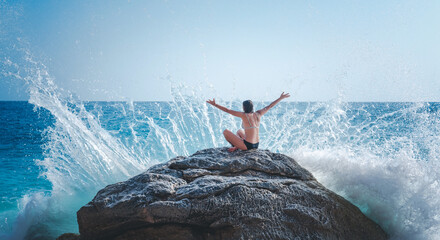 Young woman in bikin with hands up on the beach, the wave is breaking on the stone