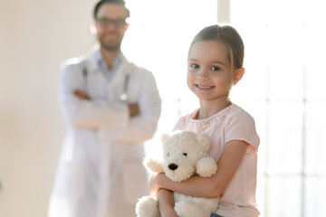 Head shot portrait smiling little girl with toy sitting in clinic with doctor pediatrician on...