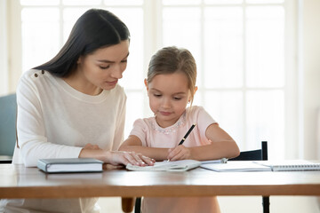Caring mother or babysitter helping little daughter with homework, learning studying at home, focused preschool girl handwriting, holding pen, sitting at work desk, homeschooling concept
