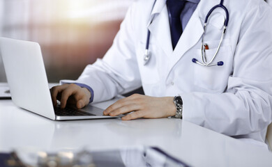 Fototapeta premium Unknown male doctor sitting and working with laptop in a darkened clinic, glare of light on the background, close-upof hands