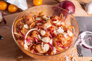 Pickled pieces of chicken with onions, garlic and pepper in a spicy chilli sauce in a glass bowl and ingredients for cooking a stew or fried dish on a wooden table. 