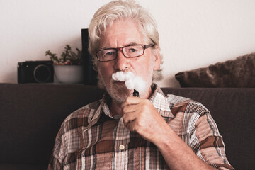 Bearded senior man smoking electronic cigarette sitting on sofa at home - new technology for more healthy smoke