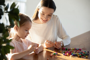 Close up loving mother helping little daughter making colorful wooden beads jewelry, sitting at...