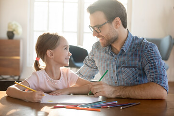 Happy father and adorable little daughter enjoying leisure time together close up, loving dad and preschool girl looking at each other, drawing colorful pencils in album, sitting at desk at home