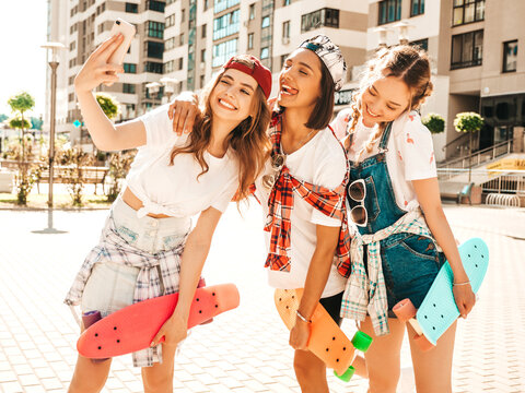 Three young smiling beautiful girls with colorful penny skateboards. Women in summer hipster clothes posing in the street background.Positive models taking selfie self portrait photos on smartphone