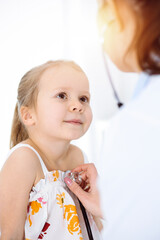 Doctor examining a child by stethoscope in sunny clinic. Happy smiling girl patient dressed in bright color dress is at usual medical inspection