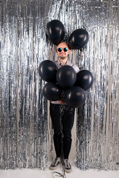 Strange caucasian man without hair in silver shirt, black leather trousers and grey trainers with black sunglasses with a lot of black baloons