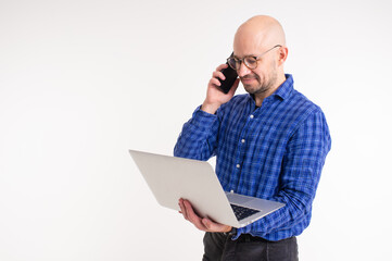 Handsome caucasian man without hair in blue shirt, black trousers and blue socks do freelance with laptop and speaks with client on phone isolated on white background