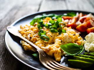Delicious breakfast - scrambled eggs with fried sausages, cottage cheese, and avocado served on black plate on wooden table
