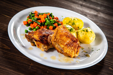 Roasted chicken thighs with boiled carrots, peas and potatoes on wooden background