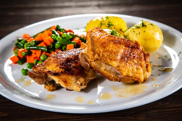 Roasted chicken thighs with boiled carrots, peas and potatoes on wooden background