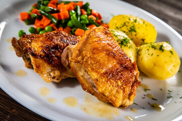 Roasted chicken thighs with boiled carrots, peas and potatoes on wooden background