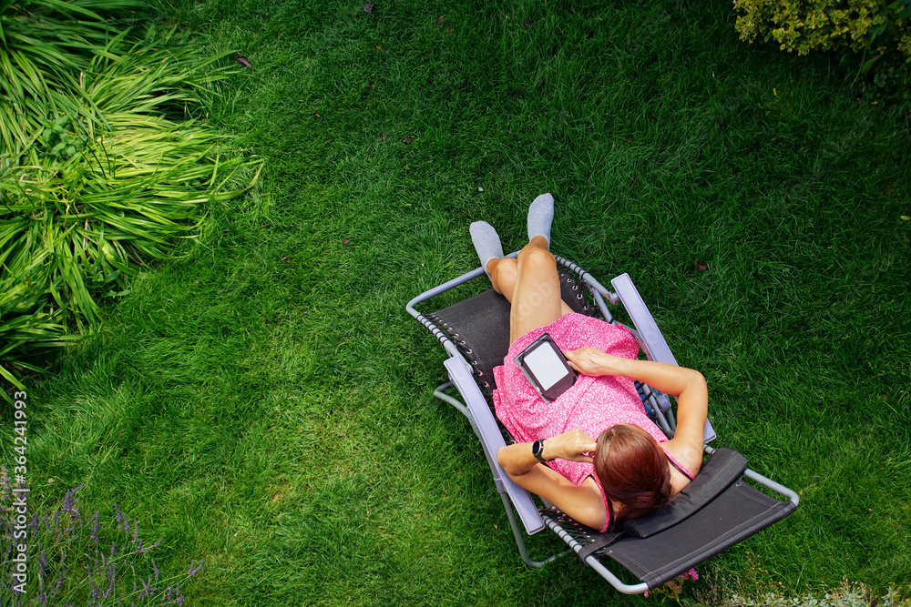 Wall mural a woman in a red dress lies on a sun lounger on the grass and reads on an ebook reader