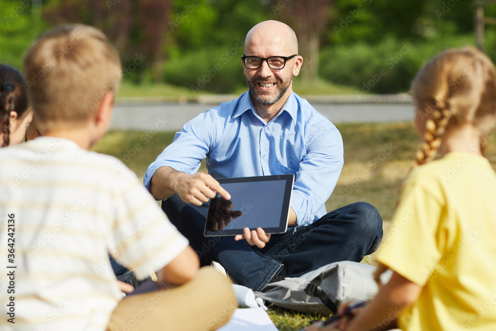 Wall mural portrait of bald male teacher pointing at tablet screen and smiling while talking to group of childr