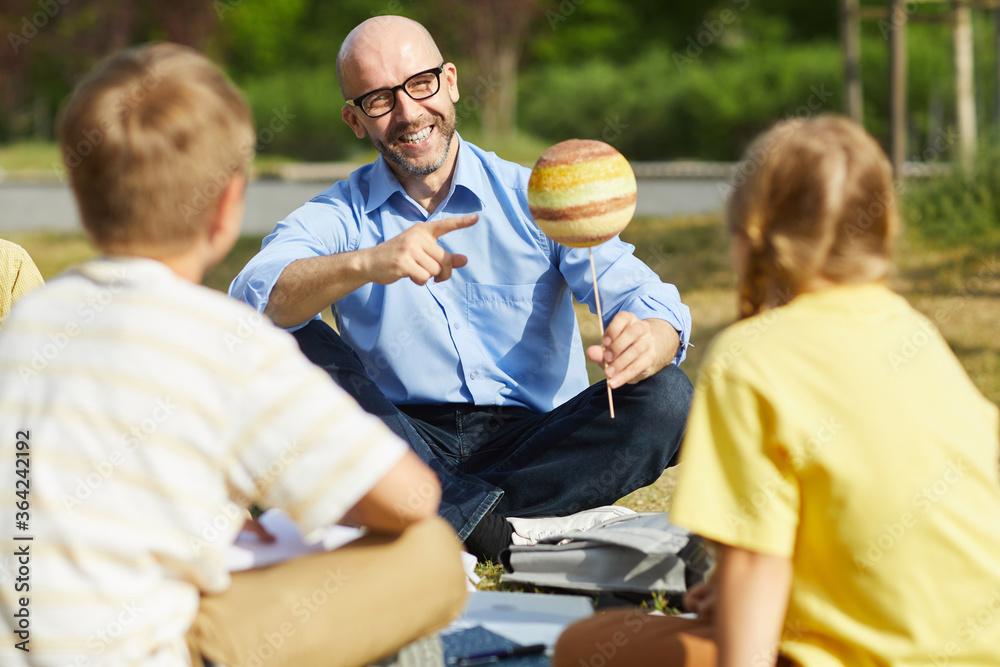 Wall mural portrait of bald male teacher pointing at planet model and smiling while talking to group of childre