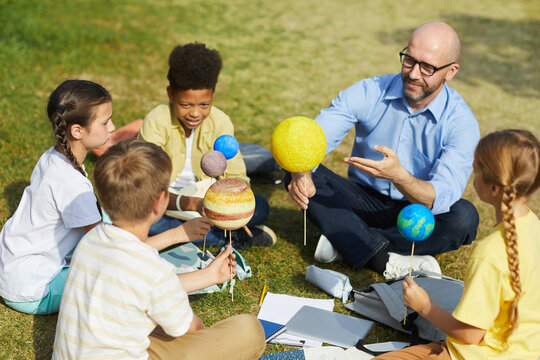 High angle portrait of male teacher pointing at planet model and smiling while enjoying outdoor astronomy class with group of children, copy space