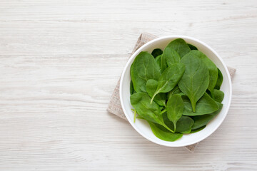 Fresh Baby Spinach in a white bowl on a white wooden background, top view. Overhead, from above, flat lay. Copy space.