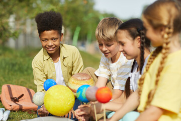 Fototapeta premium Multi-ethnic group of children sitting on green grass and holding model planets while enjoying outdoor astronomy lesson in sunlight, copy space