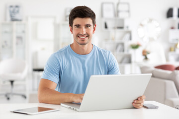 Portrait of smiling young man sitting at his desk with laptop, electronic tablet and mobile phone. Freelancer.