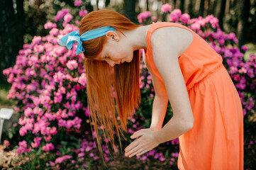 Fototapeta na wymiar Strange young girl with long red hair, pretty face in a summer orange dress cares for purple flowers