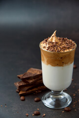 Glass of trendy fluffy creamy dalgona or whipped instant coffee with milk, coffee beans and chocolate on the dark background. New popular food and drink trend concept. Copy space.