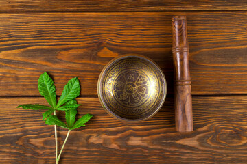 Tibetan singing bowl, wooden stick and green leaves on a wooden background. Tibetian inscription on...