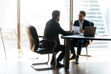 Full length focused male hr manager in glasses and formal wear holding paper resume, involved in job interview with skilled male candidate applicant, sitting together at table in modern office.